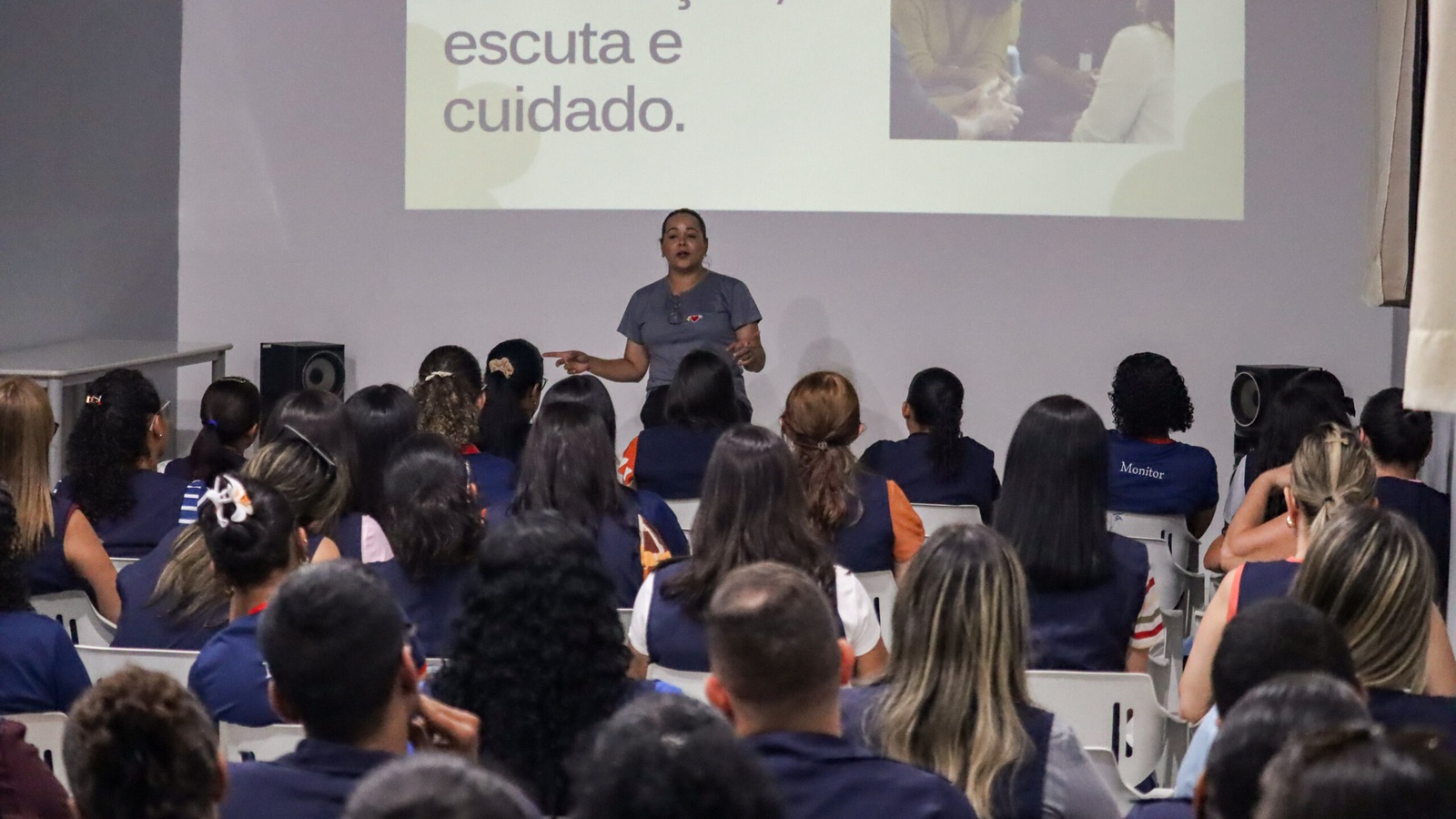 Participantes acompanham uma palestra em sala, com projeção sobre escuta e cuidado ao fundo.